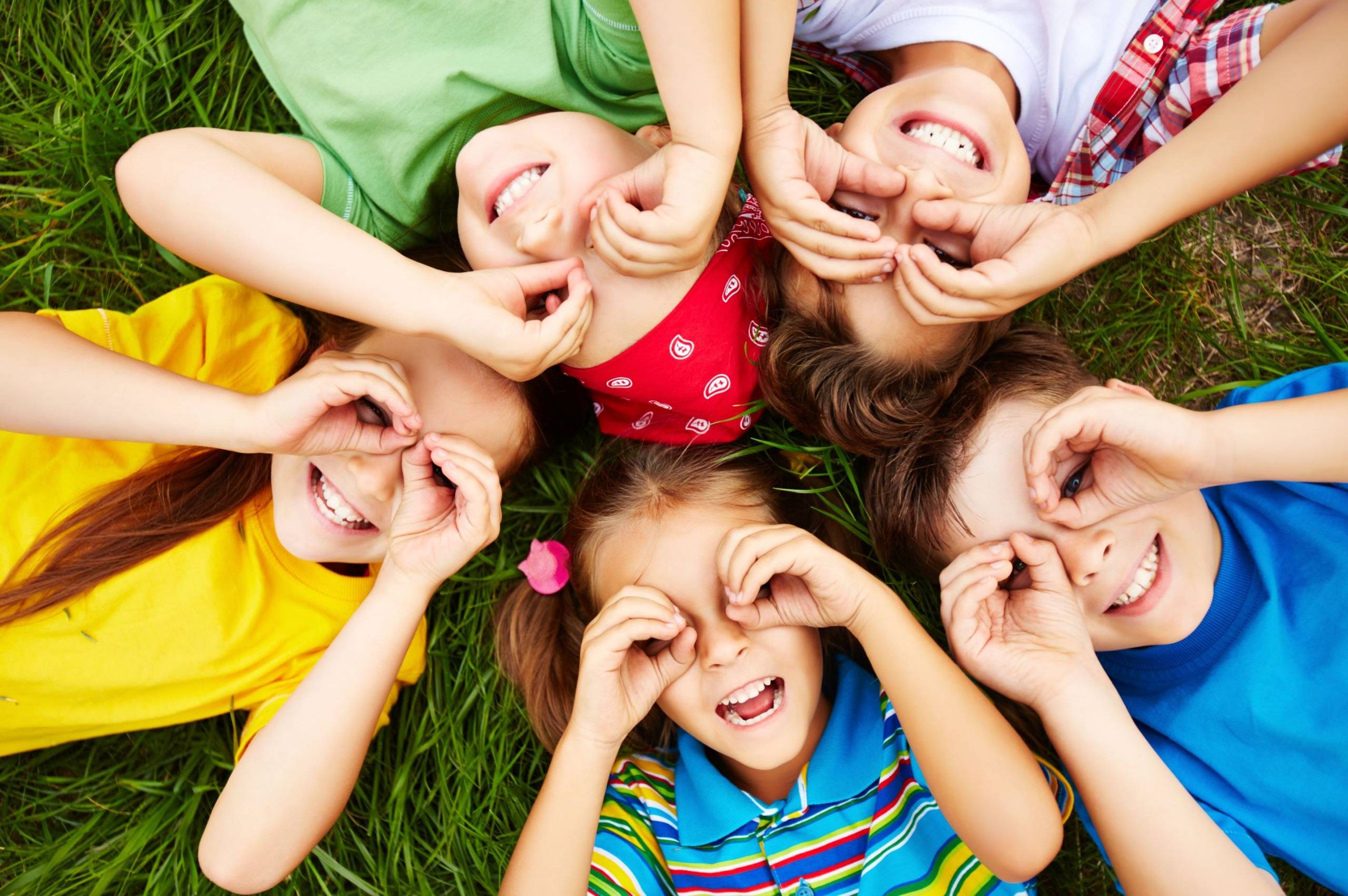 Fünf bunt gekleidete Kinder, die auf der grünen Wiese liegen und sich die Hände wie Brillen um die Augen legen.