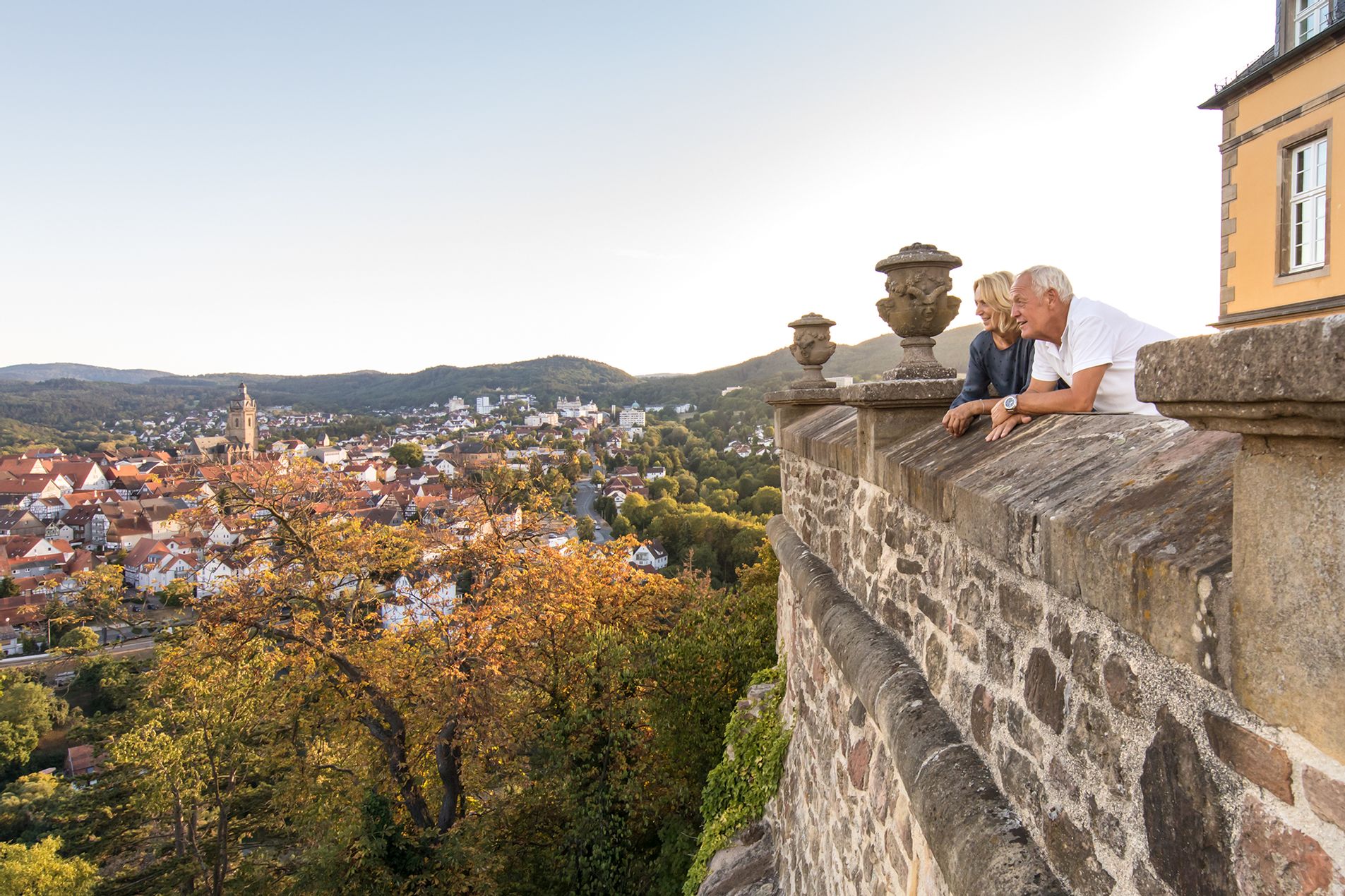 Ein Mann und eine Frau stehen rechts im Bild auf dem Schloss und schauen in die Ferne. Links im Bild ist Bad Wildungen und die Stadtkirche zu sehen.