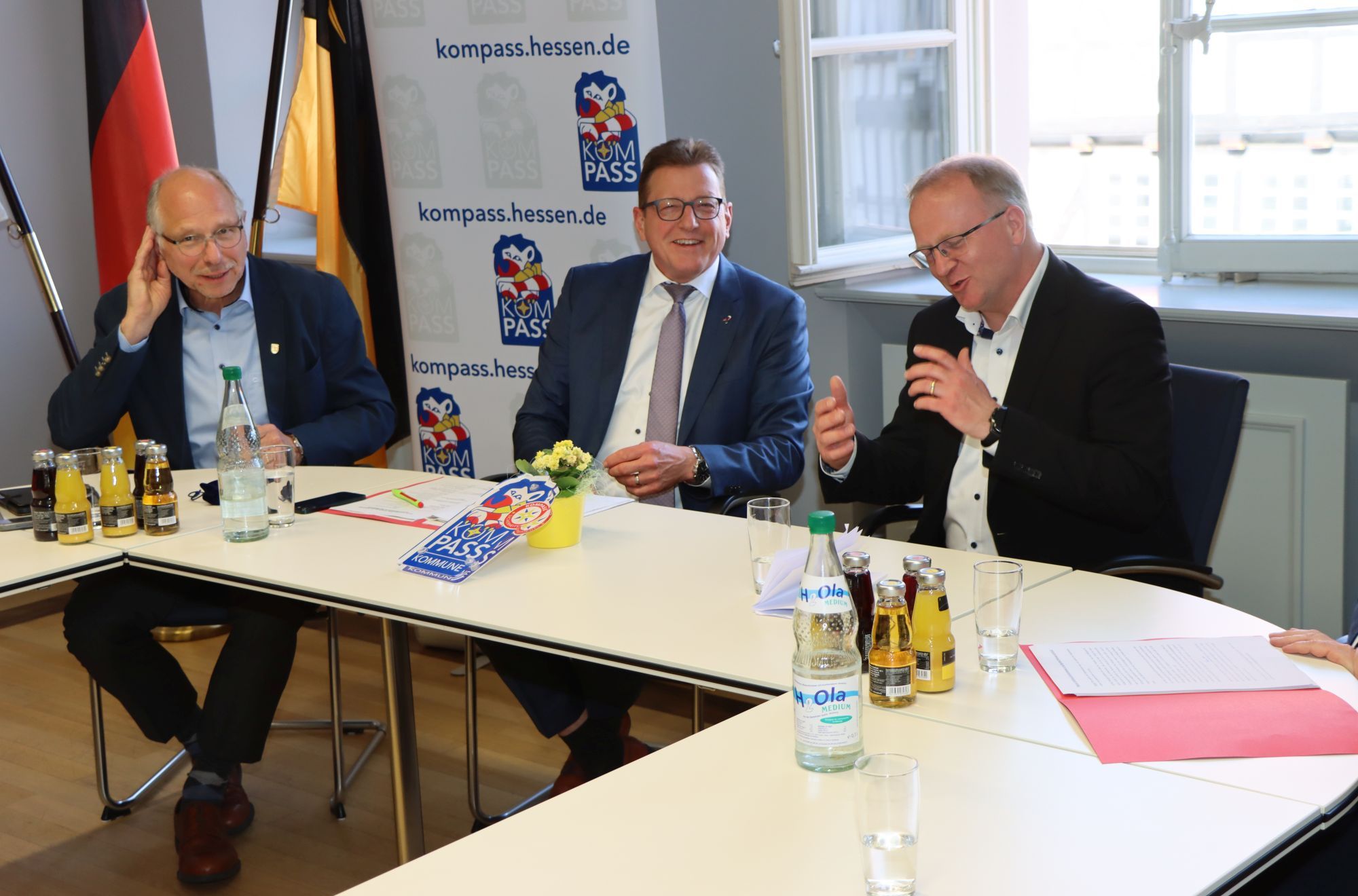 3 Beamte sitzen an einem Tisch mit dem Kompass-"Siegel" vor ihnen. Im Hintergrund stehen Banner mit der Aufschrift "kompass.hessen.de"