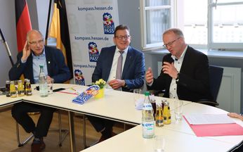 3 Beamte sitzen an einem Tisch mit dem Kompass-"Siegel" vor ihnen. Im Hintergrund stehen Banner mit der Aufschrift "kompass.hessen.de"