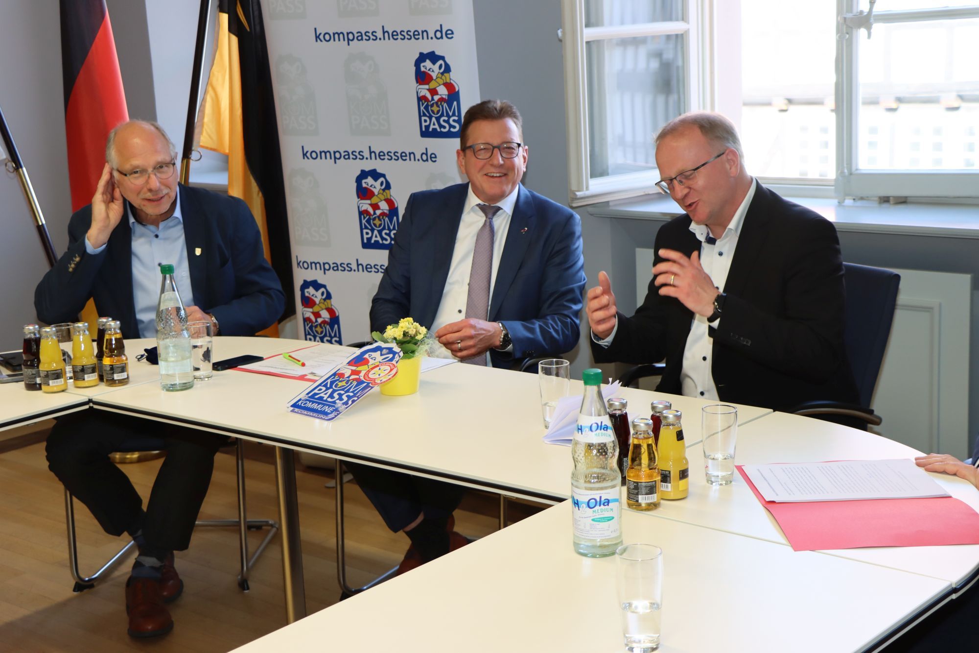 3 Beamte sitzen an einem Tisch mit dem Kompass-"Siegel" vor ihnen. Im Hintergrund stehen Banner mit der Aufschrift "kompass.hessen.de"