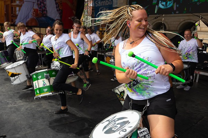 Sambaband Aipalé bei einem Auftritt. Im Vordergrund iste ine Frau zu sehen mit Trommel und grünen Sticks.