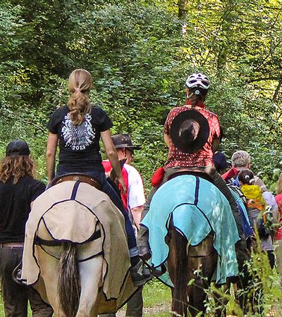 Blick von hinten auf eine Gruppe von Personen, die auf einem Feldweg läuft. 2 Personen reiten auf Pferden entlang der Gruppe