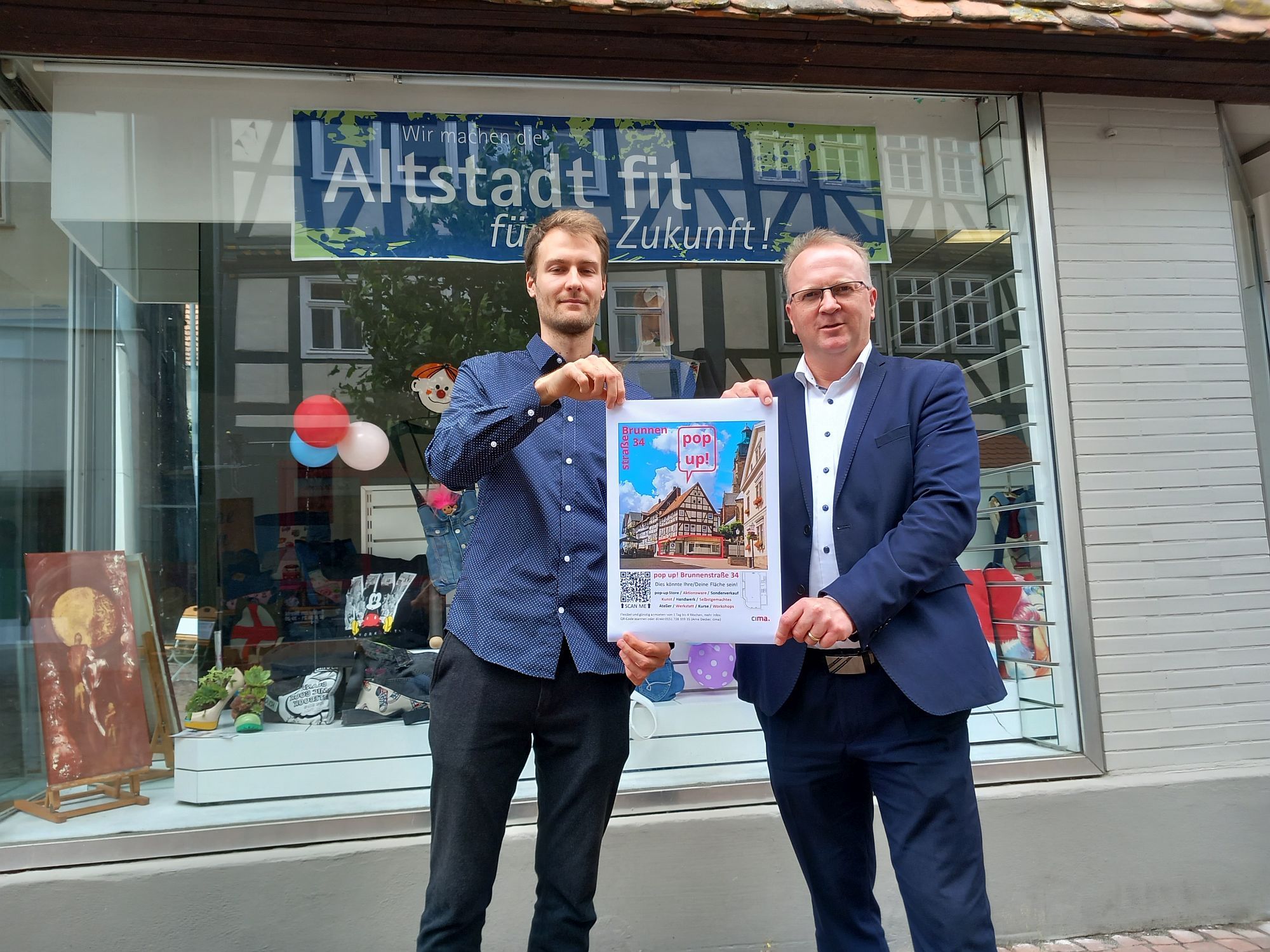 Der Bürgermeister und eine weitere Person stehen mit einem Plakat vor dem PopUp Store in der Altstadt.