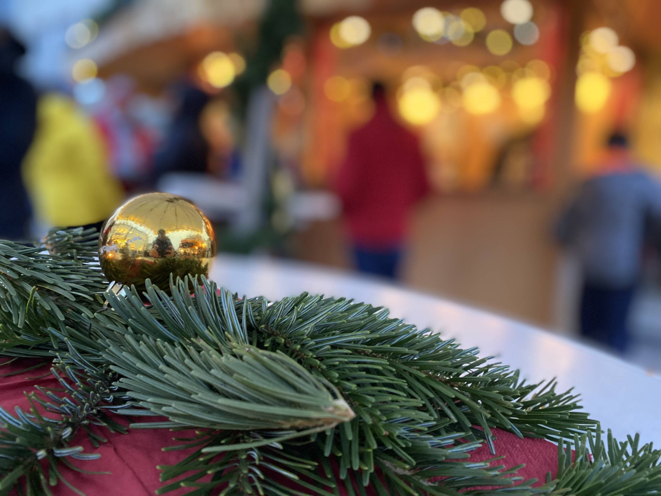 Zu sehen sind im Vordergrund in Nahaufnahme ein paar Tannenzweige und eine goldene Weihnachtskugel auf einer roten Decke liegend. Im Hintergrund verschwommen sind man Menschen an einem beleuchteten Stand auf dem Weihnachtsmarkt stehend.