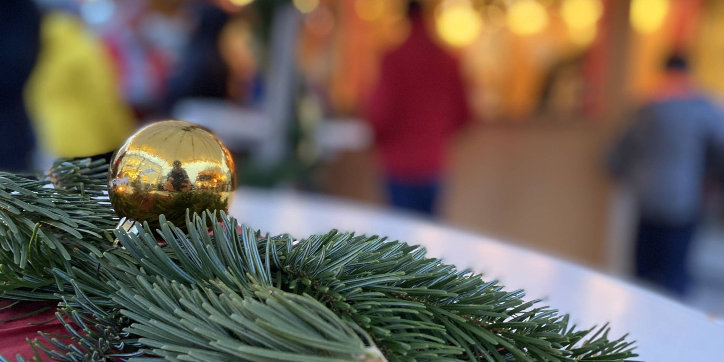 Zu sehen sind im Vordergrund in Nahaufnahme ein paar Tannenzweige und eine goldene Weihnachtskugel auf einer roten Decke liegend. Im Hintergrund verschwommen sind man Menschen an einem beleuchteten Stand auf dem Weihnachtsmarkt stehend.