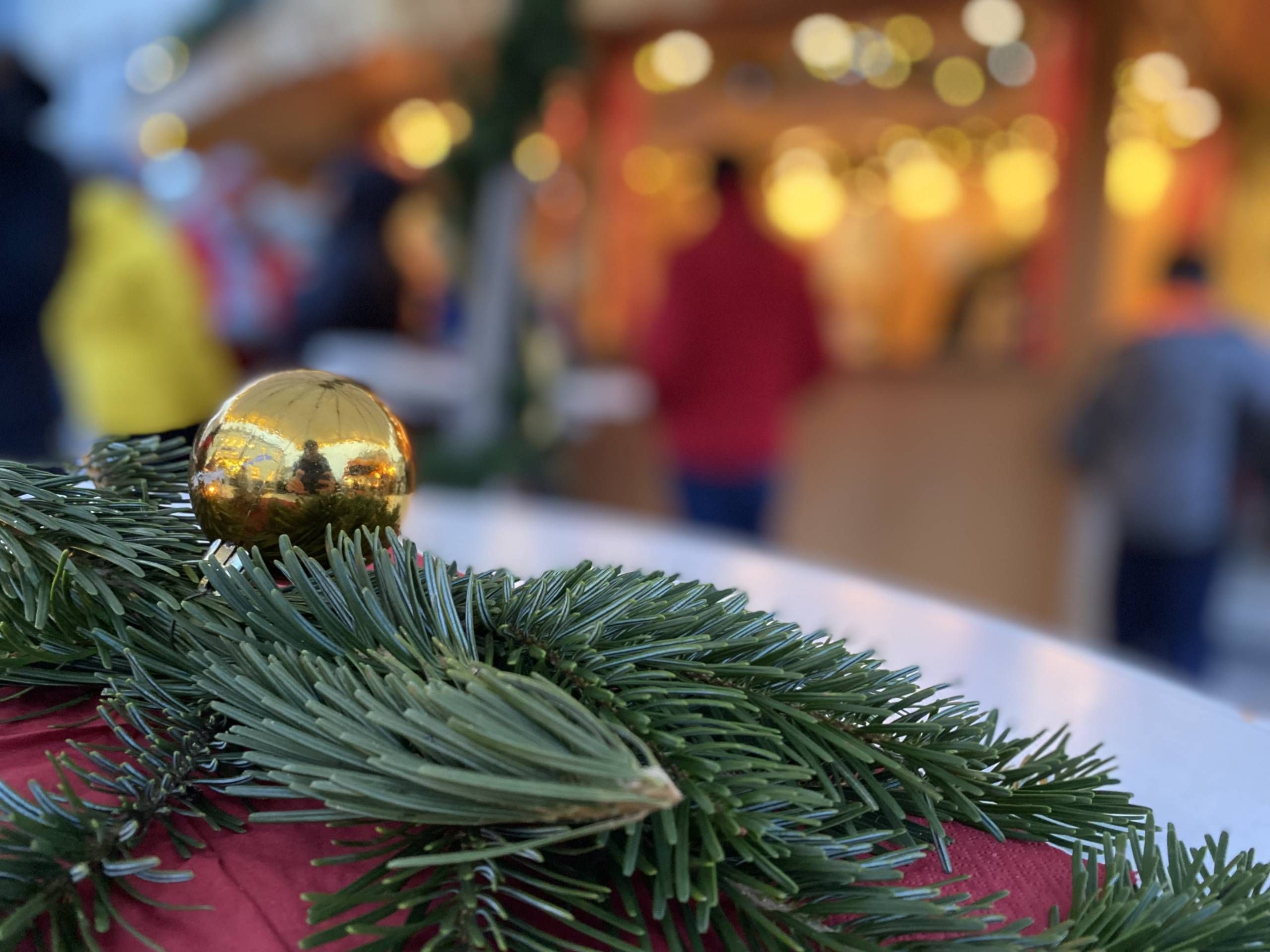 Zu sehen sind im Vordergrund in Nahaufnahme ein paar Tannenzweige und eine goldene Weihnachtskugel auf einer roten Decke liegend. Im Hintergrund verschwommen sind man Menschen an einem beleuchteten Stand auf dem Weihnachtsmarkt stehend.