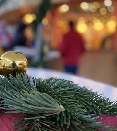 Zu sehen sind im Vordergrund in Nahaufnahme ein paar Tannenzweige und eine goldene Weihnachtskugel auf einer roten Decke liegend. Im Hintergrund verschwommen sind man Menschen an einem beleuchteten Stand auf dem Weihnachtsmarkt stehend.