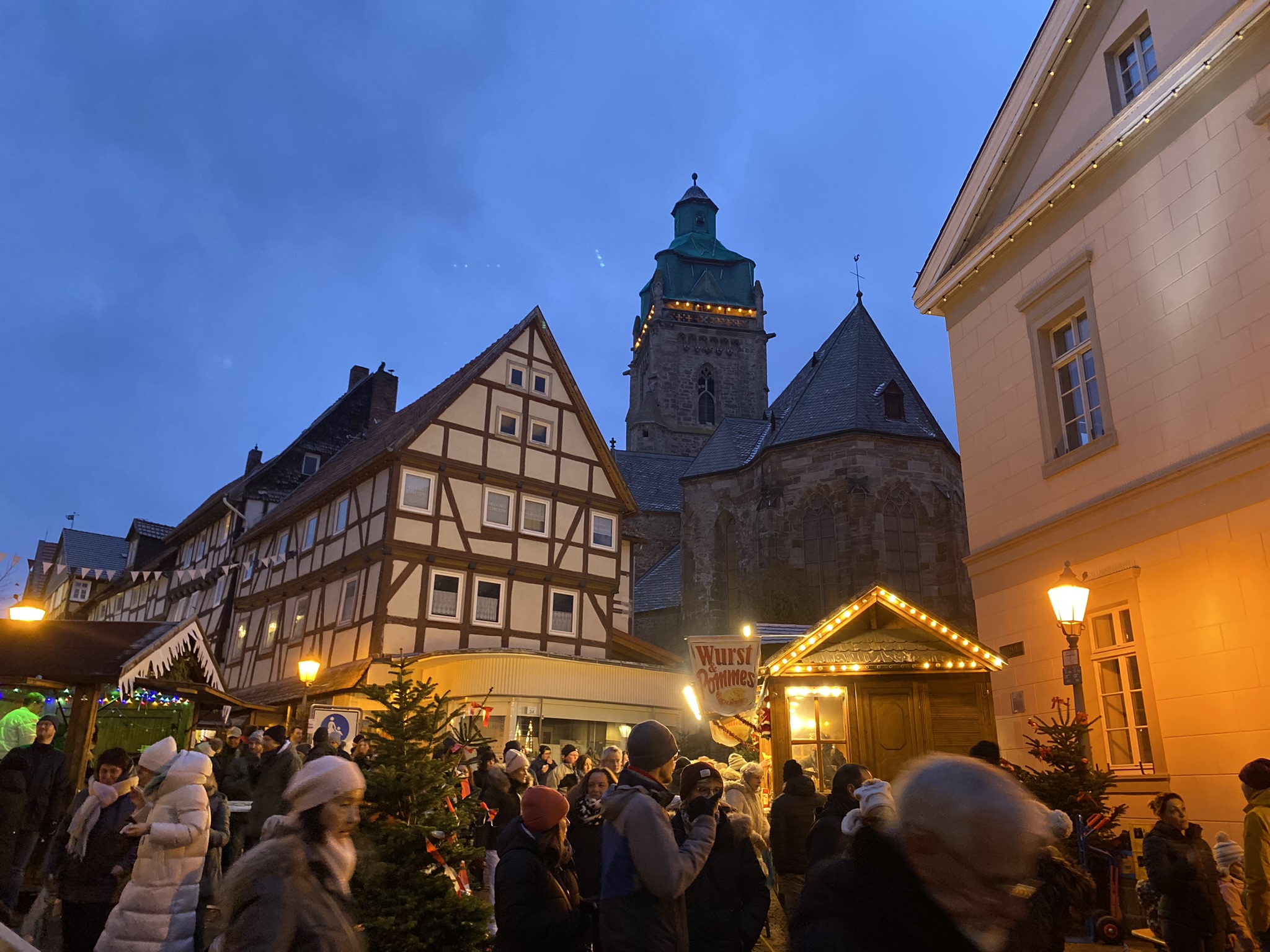 Zu sehen ist ein Teil der Bad Wildunger Altstadt-Weihnachtsmarkts am Abend. Im Hintergrund sieht man ein Fachwerkhaus, die Stadtkirche sowie einen kleinen Teil des Rathauses. Im Vordergrund sind Menschen vor Ständen zu sehen.