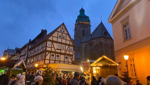 Zu sehen ist ein Teil der Bad Wildunger Altstadt-Weihnachtsmarkts am Abend. Im Hintergrund sieht man ein Fachwerkhaus, die Stadtkirche sowie einen kleinen Teil des Rathauses. Im Vordergrund sind Menschen vor Ständen zu sehen.