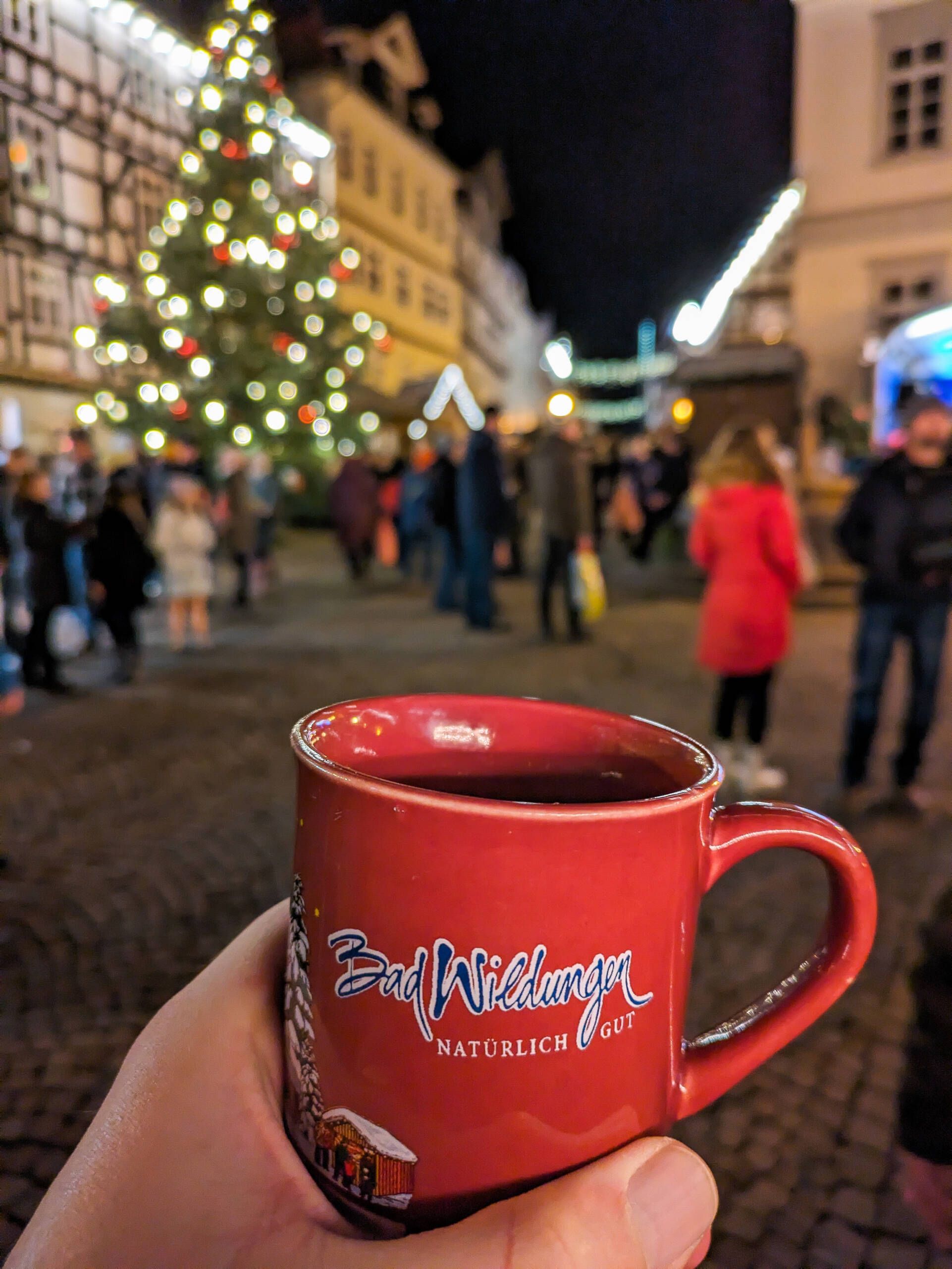 Zu sehen ist im Vordergund eine Hand die eine weihnachtliche rote Tasse hält. Im Hintergund sind Menschen und ein beleuchtetet Weihnachtsbaum zu sehen.