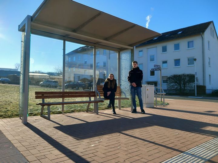 Zwei junge Personen die an einer Bushaltestelle sind und in die Kamera schauen