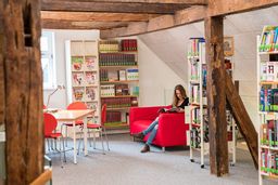 Aufnahme aus der Stadtbücherei. Eine Frau liest ein Buch auf einem Sofa zwischen Bücherregalen.