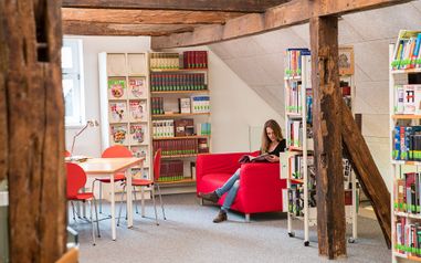 Aufnahme aus der Stadtbücherei. Eine Frau liest ein Buch auf einem Sofa zwischen Bücherregalen.