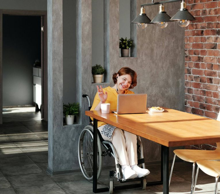 Eine Person in einem Haus, die mit einem Rollstuhl an einem Tisch sitzt und glücklich auf ihren Laptop blickt.