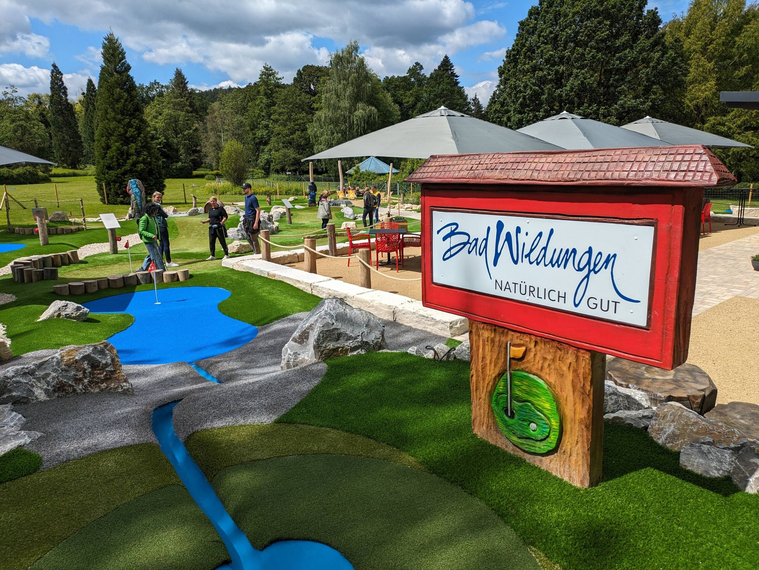 Blick auf eine Minigolfanlage. Im Vordergrund, ein Holzschild, was die Aufschrift 