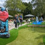 3 Personen stehen am Rand einer Minigolf Bahn und blicken auf ihre Bälle, die in der Nähe des Lochs liegen.