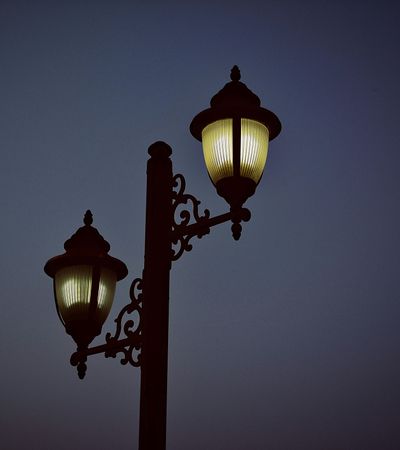 Eine altmodische Straßenlaterne mit zwei Laternenköpfen die vor einem dunkelblauen Nachthimmel leuchtet.