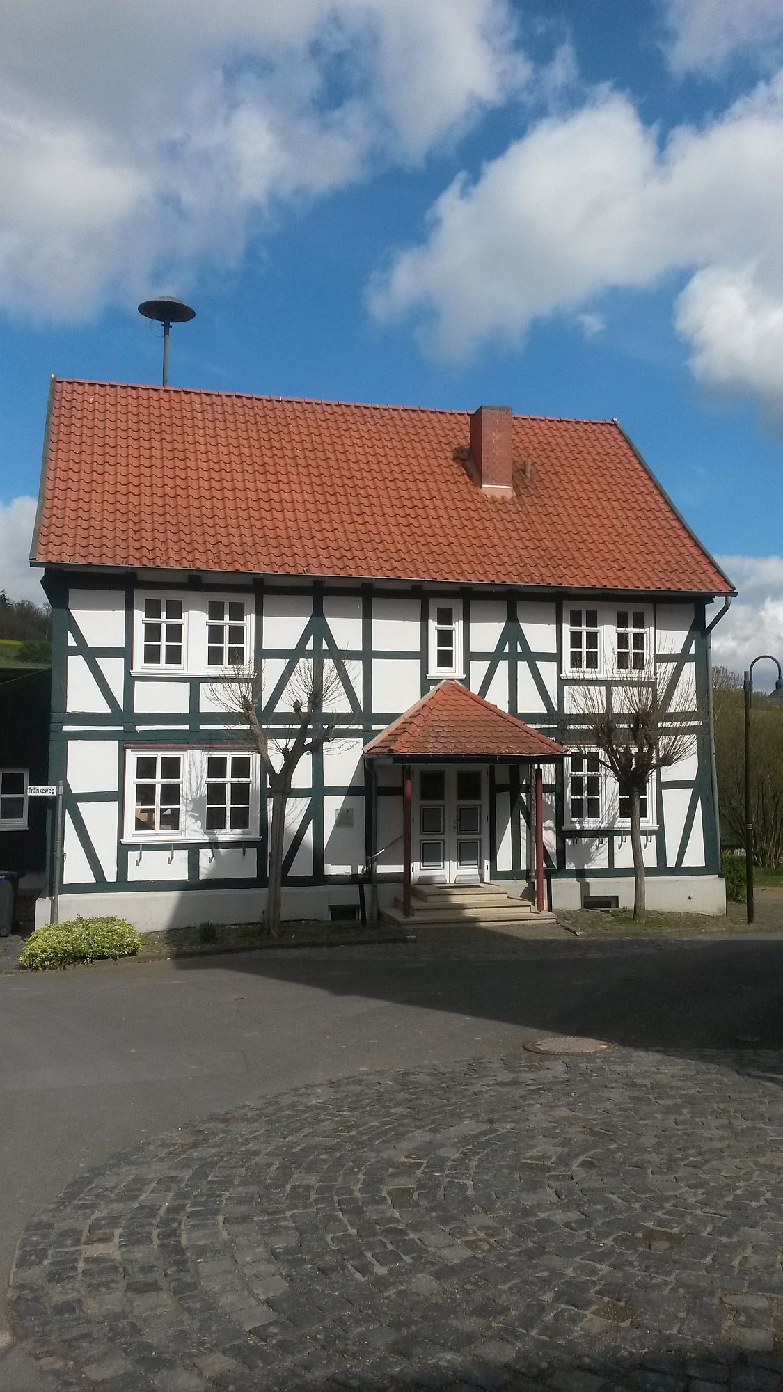 Ein grün weißes Fachwerk Haus. Der Himmel ist blau und bewölkt. Vor dem Haus stehen 2 kahle Bäume.