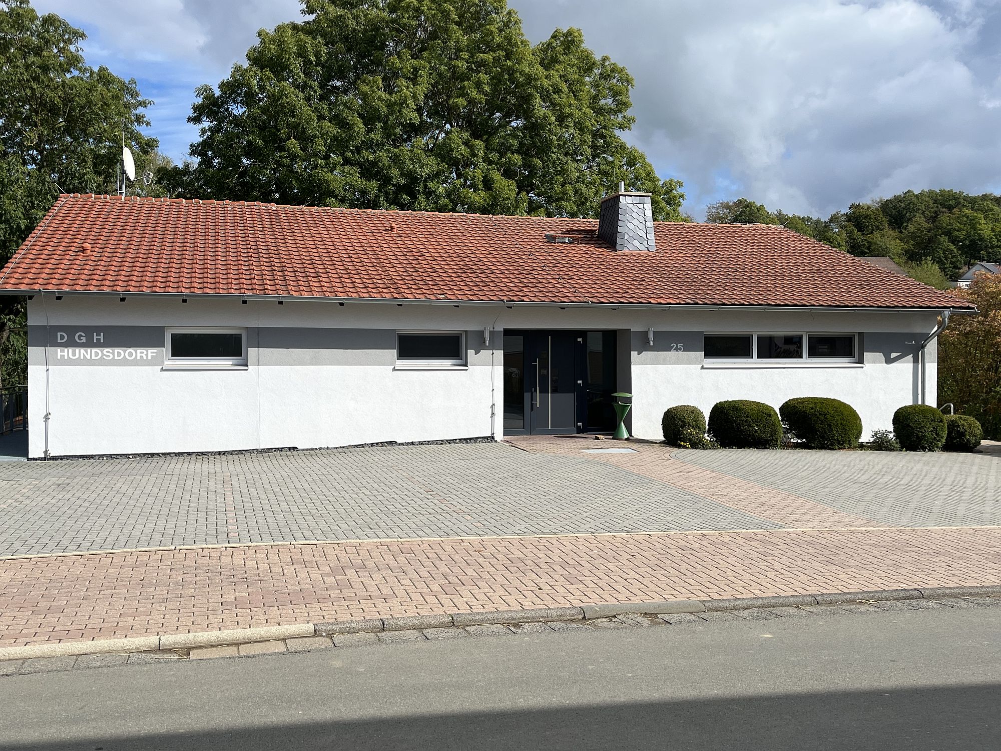 Außenaufnahme von der Frontansicht des Dorfgemeinschaft Haus Hundsdorf. Zu sehen sind leere Parkplätze und Bäume im Hintergrund.