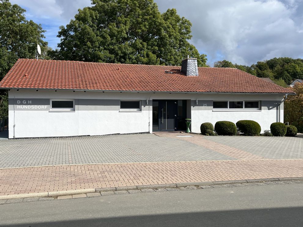 Außenaufnahme von der Frontansicht des Dorfgemeinschaft Haus Hundsdorf. Zu sehen sind leere Parkplätze und Bäume im Hintergrund.