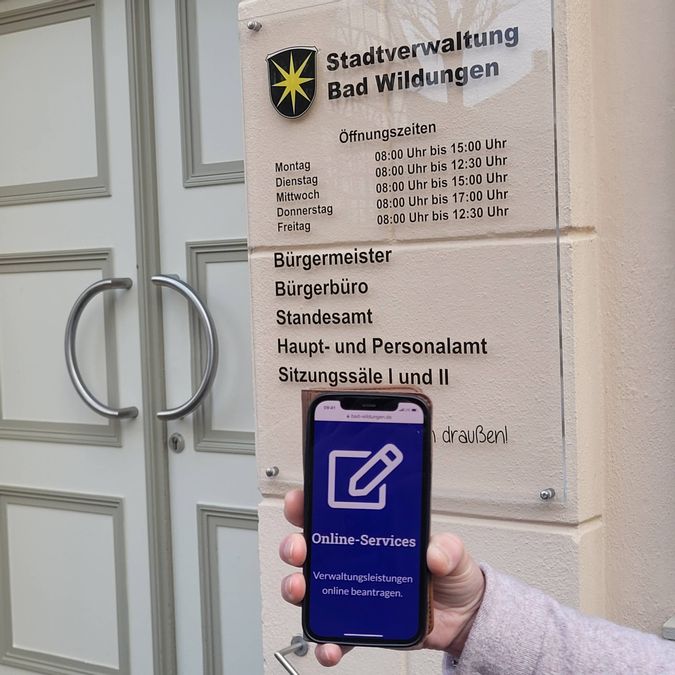 Die Eingangstür des Rathauses mit einem Schild der Öffnungszeiten ist zu sehen. Davor eine Hand, die ein Handy hält mit dem Text "Online Services"