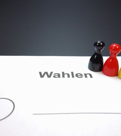 3 verschieden farbige Spielfiguren stehen auf einem Blatt Papier, das mit "Wahlen" beschriftet ist.