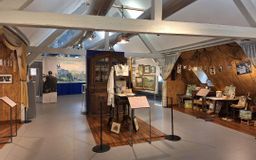 Auf dem Bild ist ein Teil der neuen Dauerausstellung von Willi Tillmans zu sehen: Einige seiner Werke sowie ein Teil seines Ateliers mit Tisch, Stühlen, Schrank und aufgehängten Bildern.