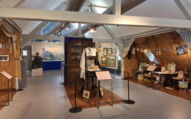 Auf dem Bild ist ein Teil der neuen Dauerausstellung von Willi Tillmans zu sehen: Einige seiner Werke sowie ein Teil seines Ateliers mit Tisch, Stühlen, Schrank und aufgehängten Bildern.