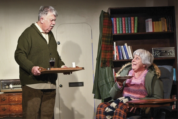 Zu sehen ist ein älterer Mann mit einem Tablett in der Hand, der vor einer Frau in einem Sessel steht, die ein Teller in der Hand hält. Im Hintergrund ist eine Tür sowie in Bücherregal zu sehen.