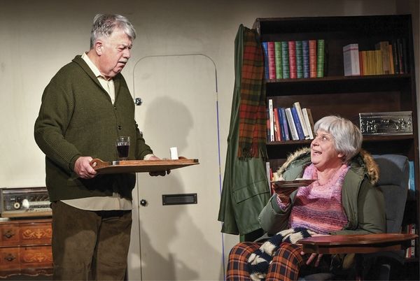 Zu sehen ist ein älterer Mann mit einem Tablett in der Hand, der vor einer Frau in einem Sessel steht, die ein Teller in der Hand hält. Im Hintergrund ist eine Tür sowie in Bücherregal zu sehen.