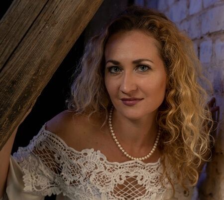 Zu sehen ist die Pianistin Vera Osina in einem weißen Kleid an einer Mauer stehend schaut sie unter einem Holzbalken durch.