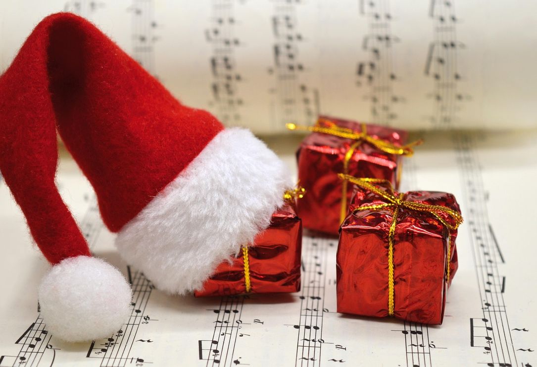 Zu sehen sind drei kleine rote Geschenke mit goldenem Band auf einem Notenheft liegend. Eines dieser Geschenke hat eine rot-weiße Weihnachtsmannmütze auf.