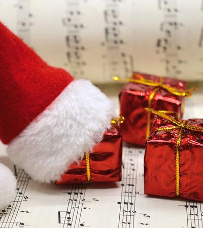 Zu sehen sind drei kleine rote Geschenke mit goldenem Band auf einem Notenheft liegend. Eines dieser Geschenke hat eine rot-weiße Weihnachtsmannmütze auf.