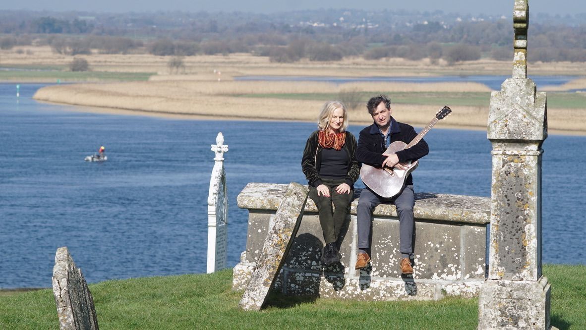 Zu sehen sind die beiden Musiker Eleanor Shanley & Ultan Conlon auf einer Mauer sitzend. Im Hintergrund ist ein Gewässer sowie eine Küste zu sehen.