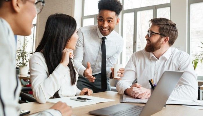 Vier gut gelaunte Personen sitzen oder stehen um einem Schreibtisch herum und diskutieren