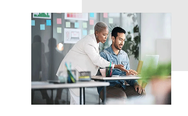 In a professional setting, a man and a woman are discussing employee feedback. The man is sitting at a table and showing his colleague management feedback from his laptop.