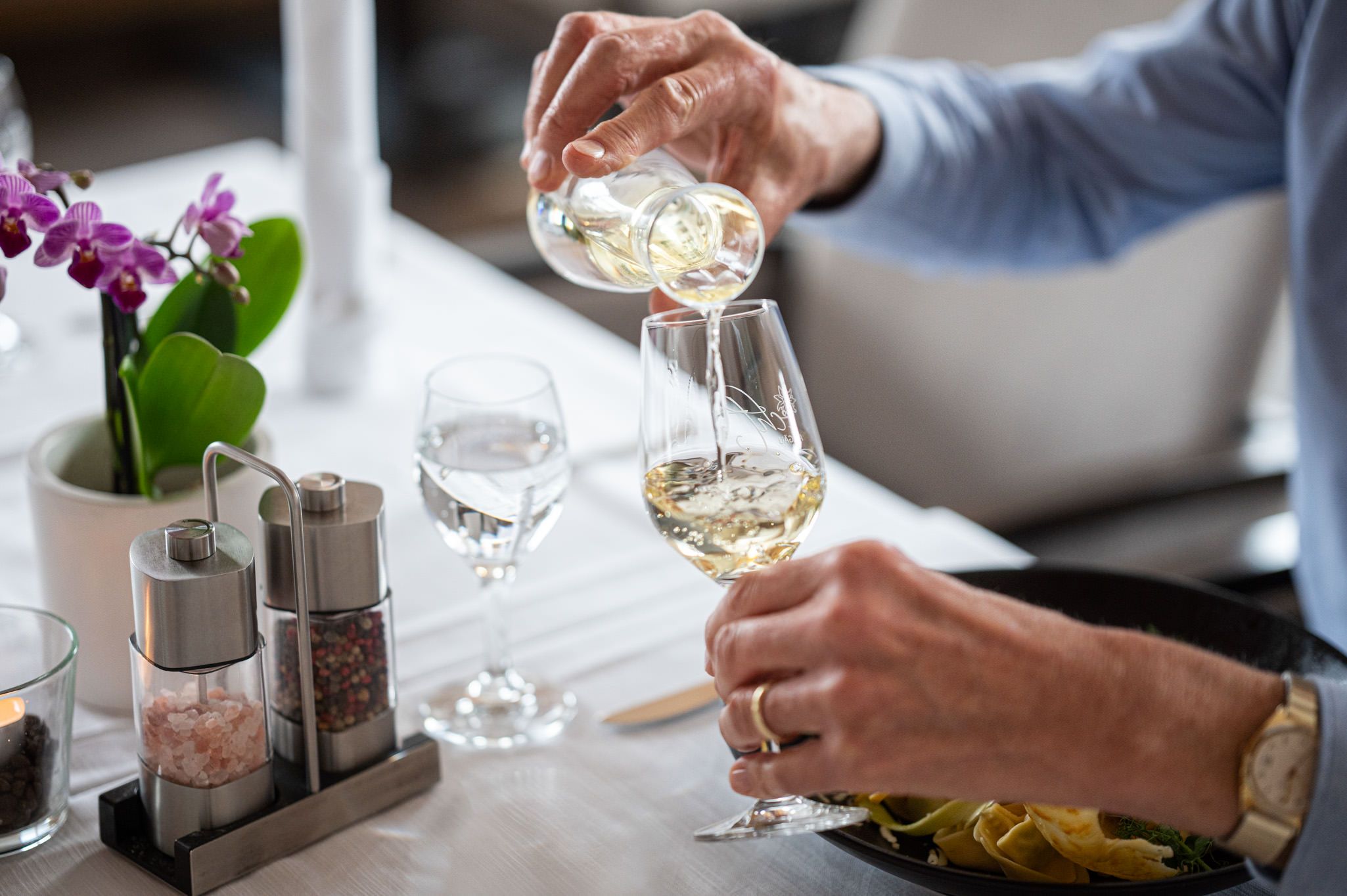 Nahaufnahme einer Hand, die an einem stilvoll gedeckten Tisch ein Glas Rotwein in ein Weinglas einschenkt – ein Moment genussvoller Gastlichkeit in der Speisegalerie Fischen, wo kulinarische Vielfalt und kulturelles Ambiente aufeinandertreffen.