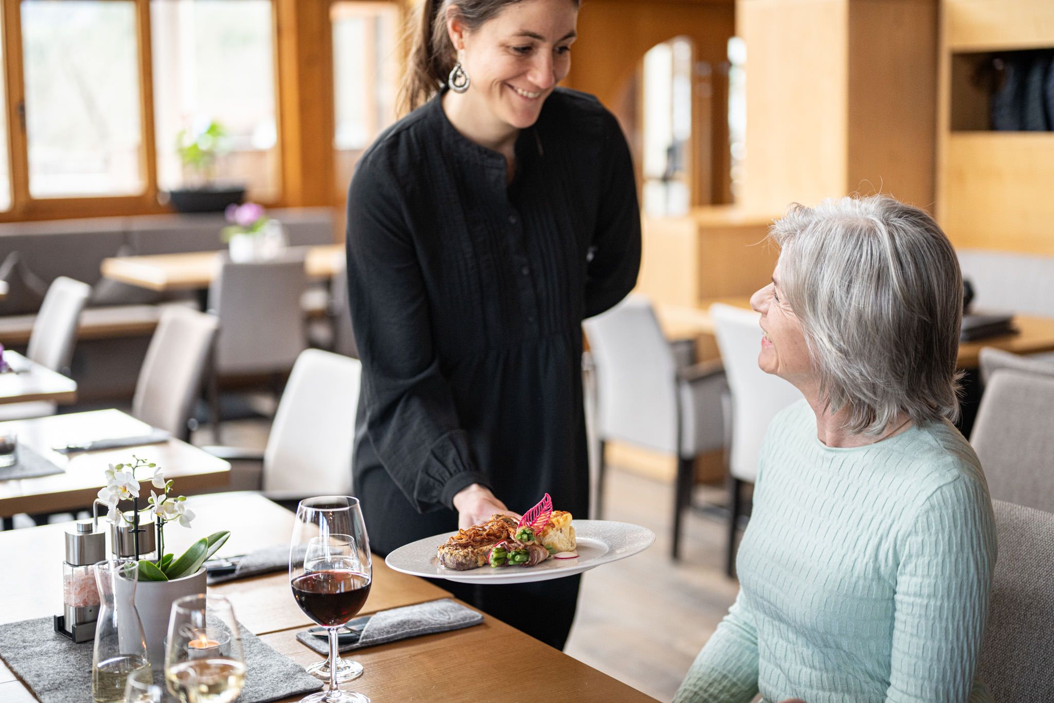 Eine Kellnerin serviert einer Frau ihr Essen, während diese ein Glas Rotwein genießt. Beide lächeln sich freundlich an – ein Moment herzlicher Gastlichkeit in der Speisegalerie Fischen, wo Genuss und persönliche Atmosphäre im Mittelpunkt stehen.