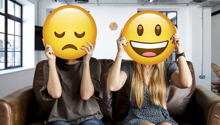2 Mitarbeiterinnen im Büro halten sich Smileys vors Gesicht: Gutes Betriebsklima und schlechtes Betriebsklima.