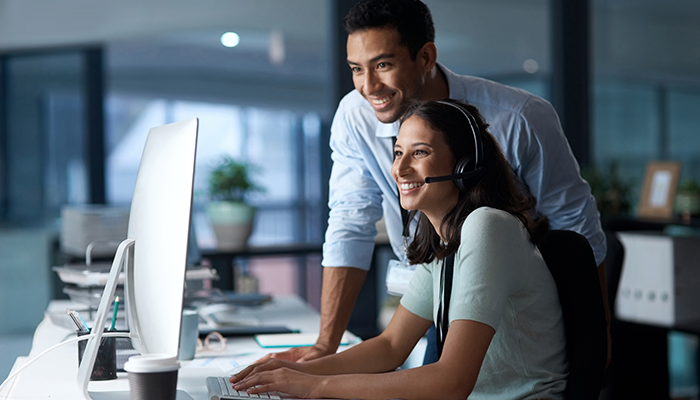 Eine Frau sitzt mit einem Headset am Computer und ihr Kollege unterstützt sie bei einer Community-Software.