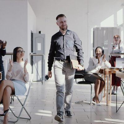 Einen einem positiven Führungxskräftefeedback läuft ein Mann mit Unterlagen zufrieden durch das Büro und wird dabei von seinen Kollegen und Kolleginnen positiv empfangen.