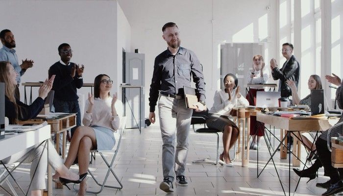 Einen einem positiven Führungxskräftefeedback läuft ein Mann mit Unterlagen zufrieden durch das Büro und wird dabei von seinen Kollegen und Kolleginnen positiv empfangen.