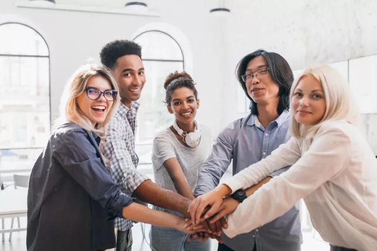 Junges multinationales Team symbolisiert soziale Gleichheit in dem es in die Kamera lächelt und als Ausdruck des Zusammenhalts die Hände aufeinander hält