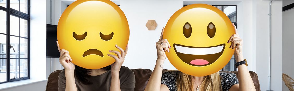 2 Mitarbeiterinnen im Büro halten sich Smileys vors Gesicht: Gutes Betriebsklima und schlechtes Betriebsklima.