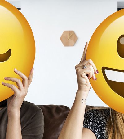 2 Mitarbeiterinnen im Büro halten sich Smileys vors Gesicht: Gutes Betriebsklima und schlechtes Betriebsklima.