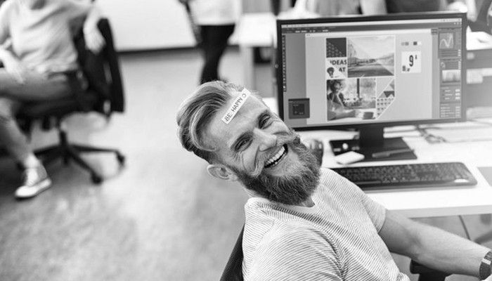 Ein lachender Mann am Arbeitsplatz mit "Be Happy"-Zettel auf der Stirn steht symbolisch für ein gutes Betriebsklima.