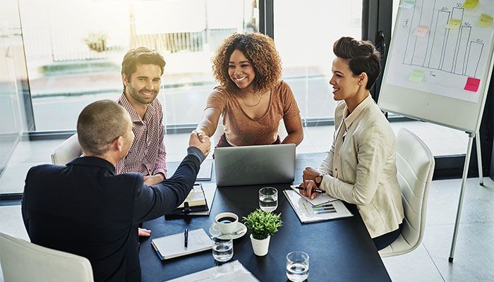 Vier Menschen sitzen an einem Tisch und zwei geben sich dabei die Hand, weil sie vereinbart haben eine Feedbackautomatisierung Ihres Führungskräftefeedbacks durchzuführen.
