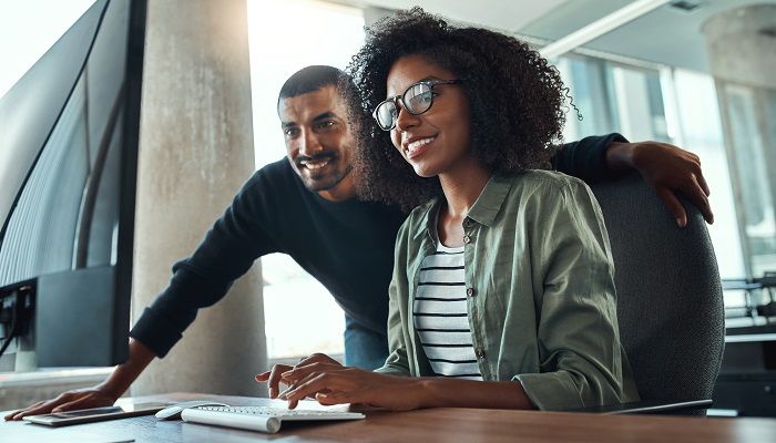 An african young businesswoman sitting at her desk working on computer with male colleague standing by