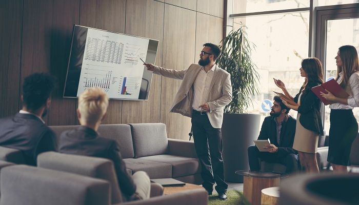 Ein Manager gibt seinen Mitarbeitern Führungskräftefeedback und zeigt dabei auf Daten auf einem Bildschirm.